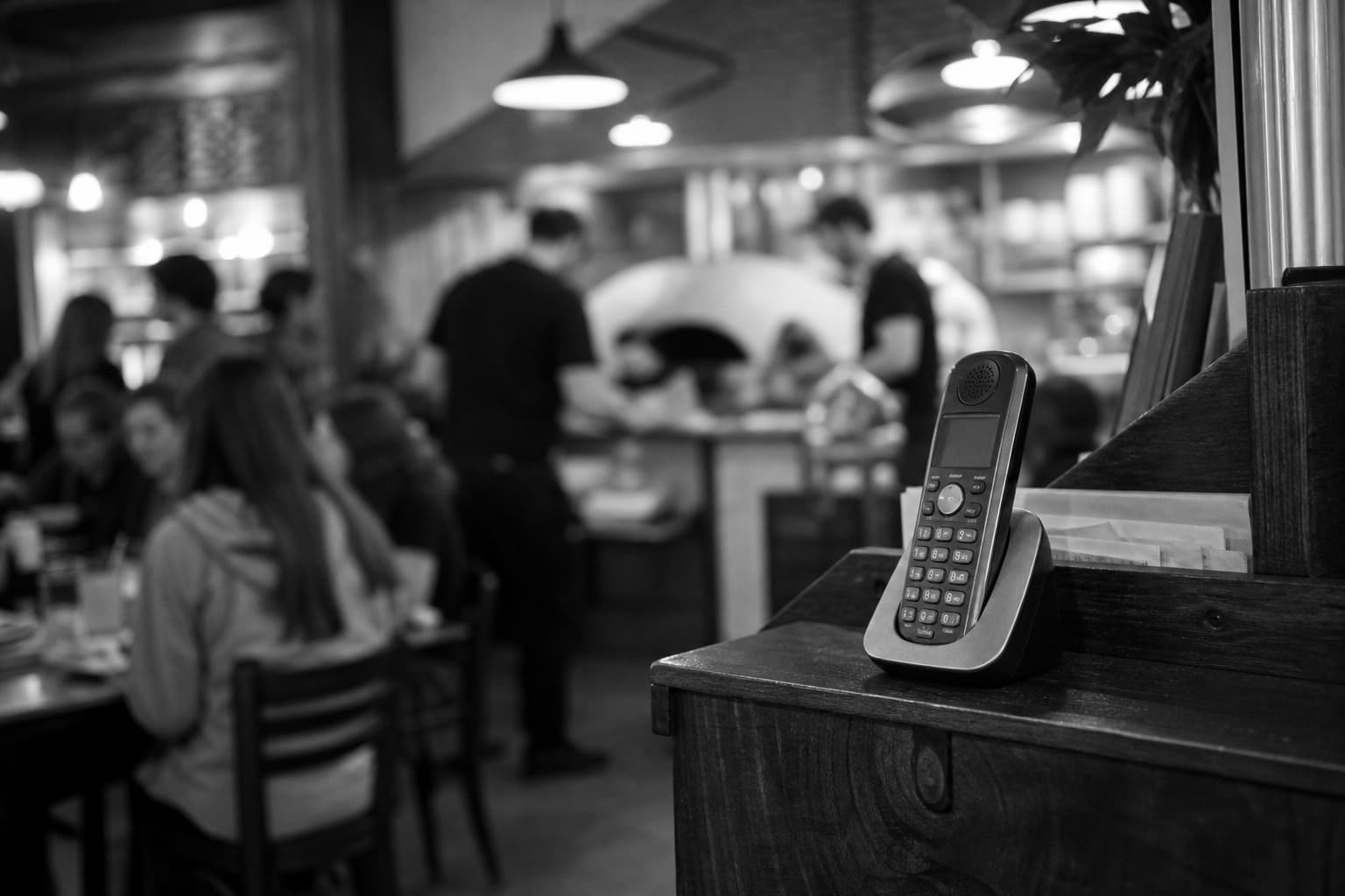 A cordless phone in its cradle on a wooden host stand, with a busy pizza shop blurred behind it